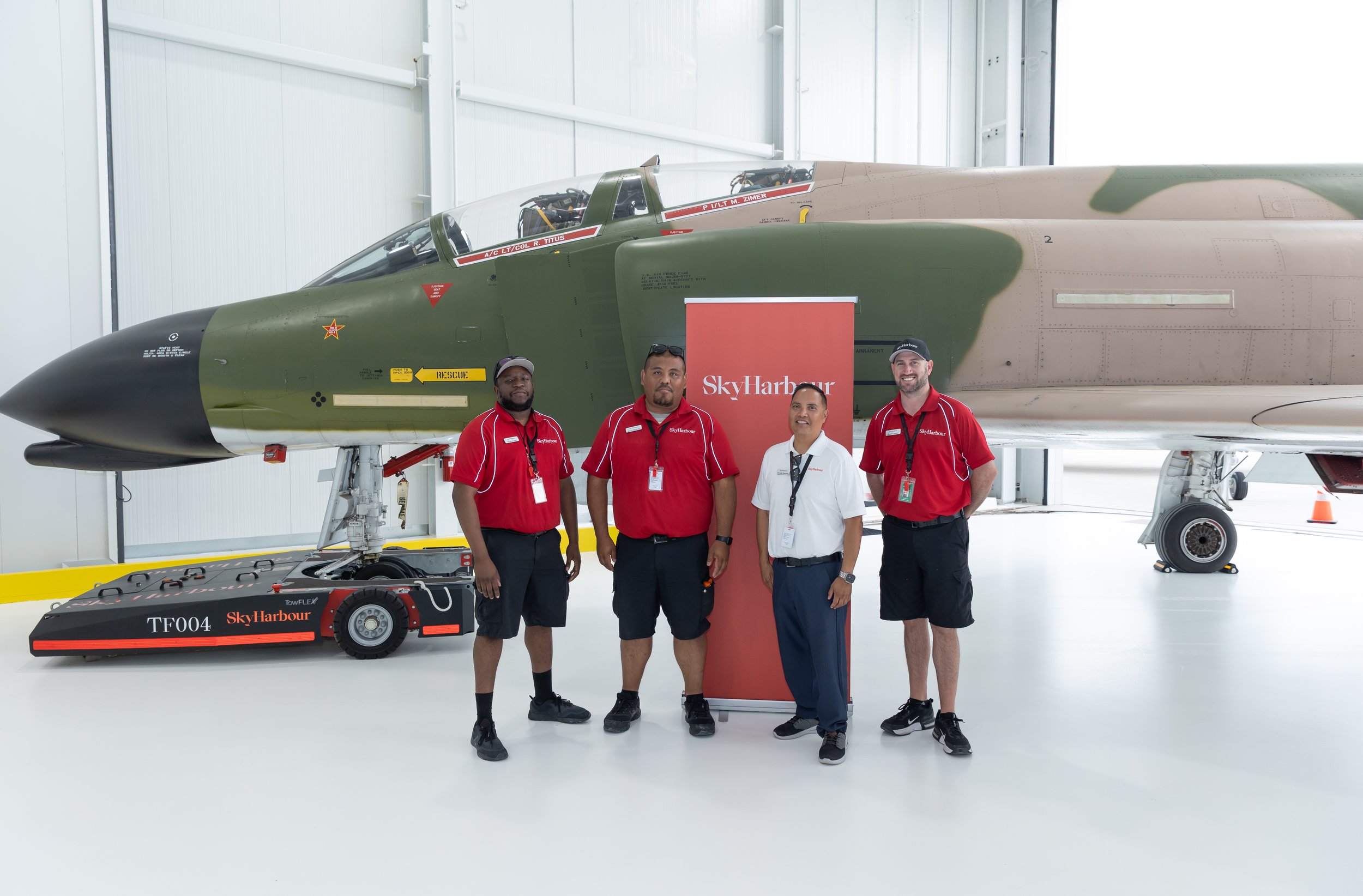 Four people in red uniform shirts standing beside a vintage military jet inside a hangar