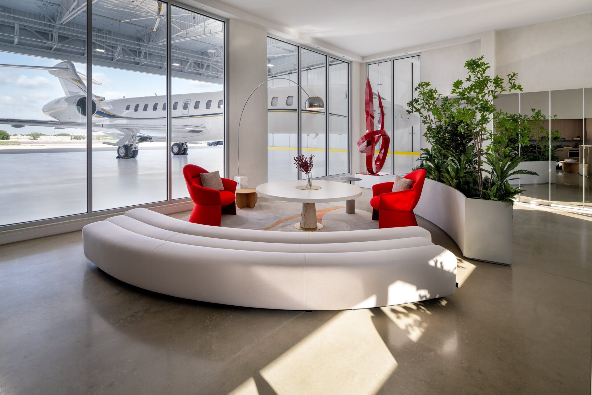 Hangar lounge with a curved white sofa, red chairs and floor-to-ceiling windows overlooking a private jet