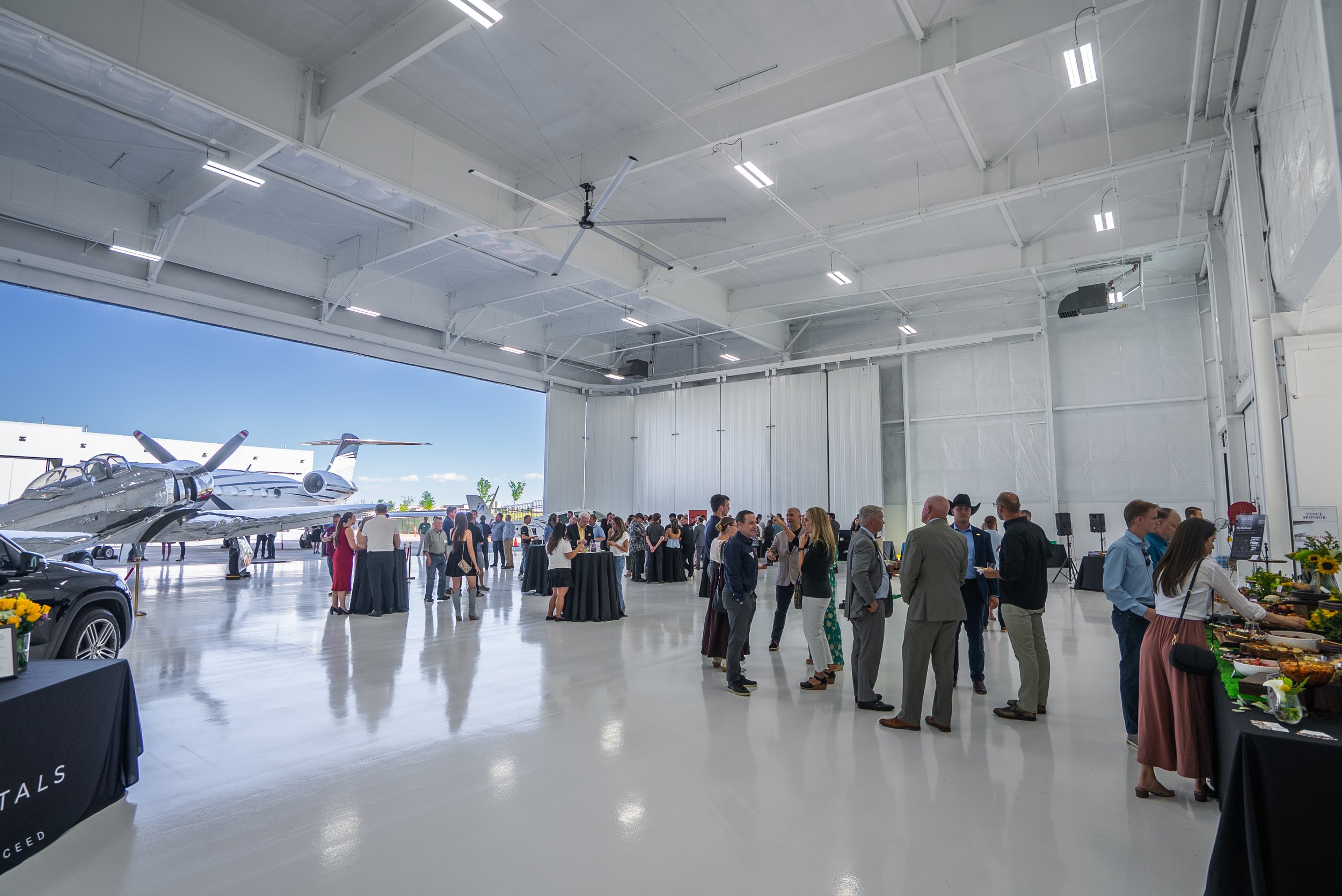 Crowd of guests gathered inside a hangar at a reception, with a jet visible through the open door