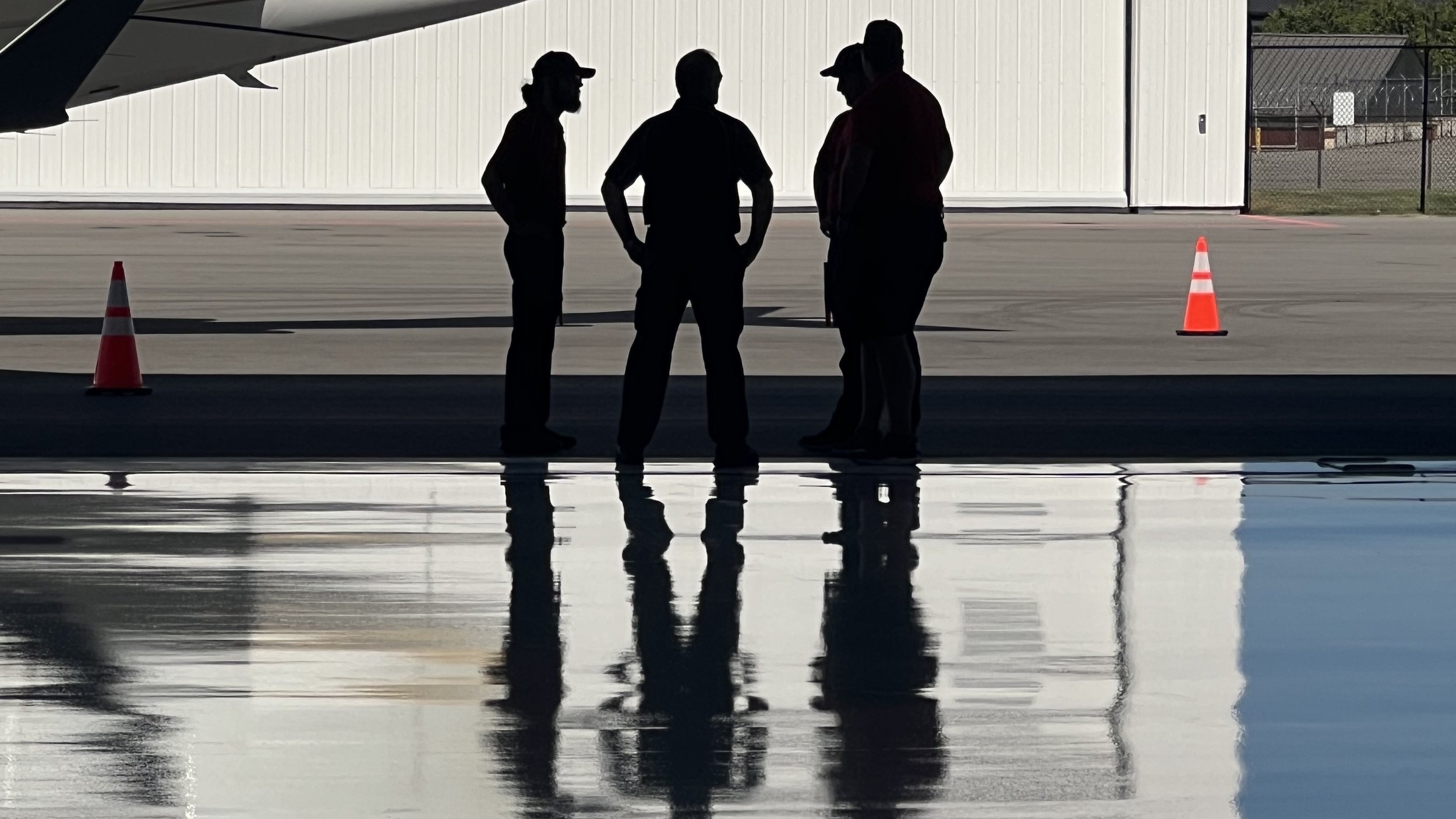Three silhouetted people standing in a hangar doorway with reflections on the floor