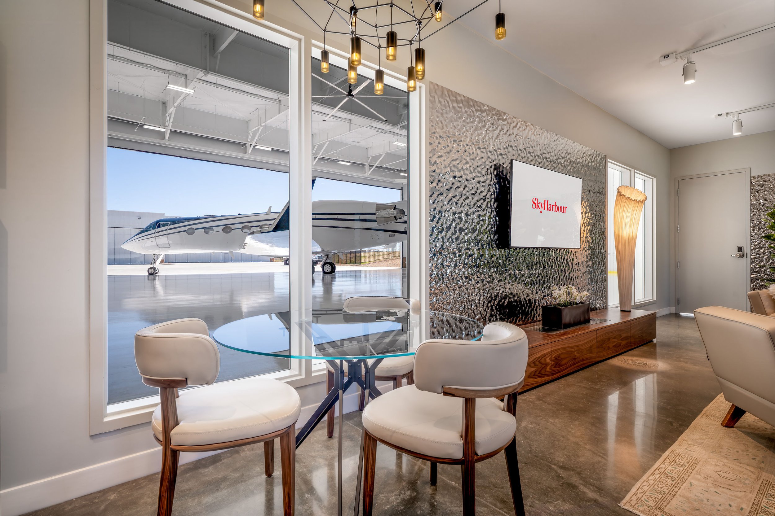 Hangar lounge with cream chairs and a glass table beside a window overlooking the ramp and a jet