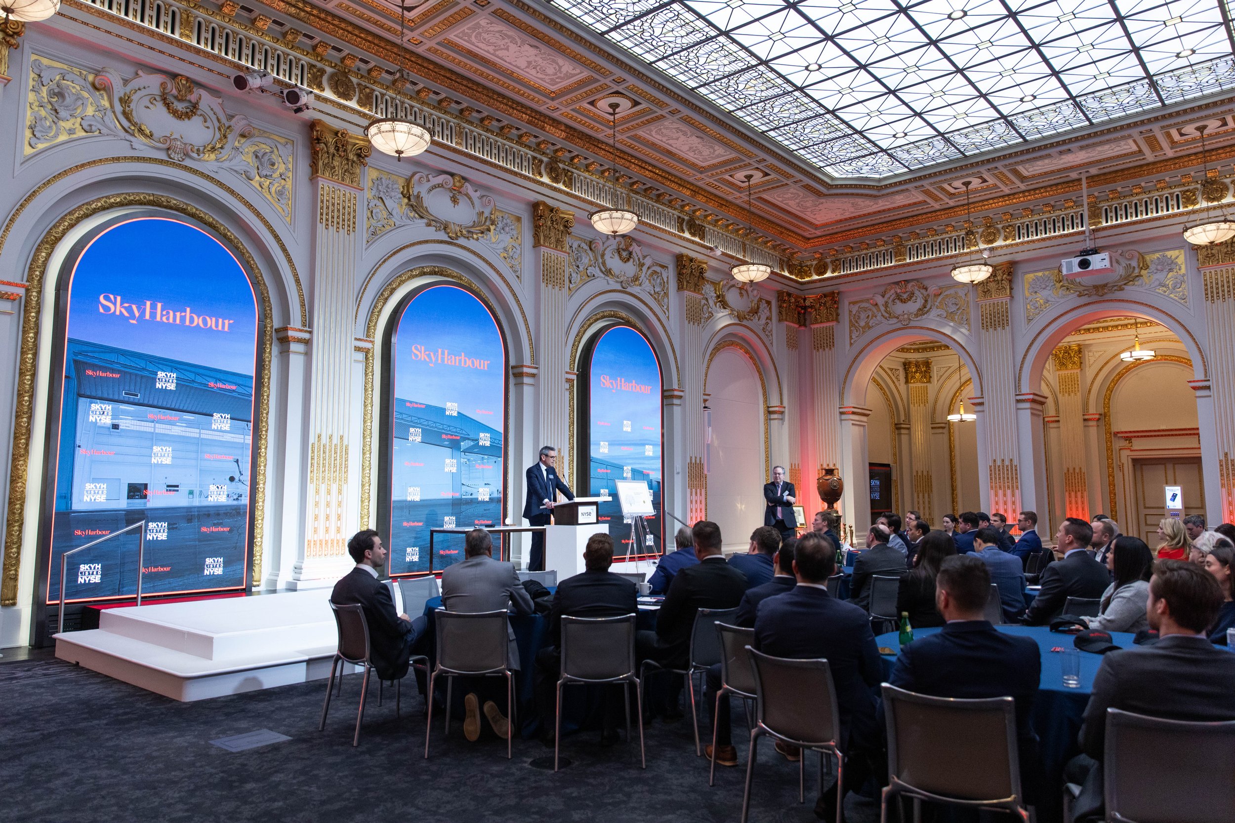 Audience seated in an ornate hall with arched windows, watching a speaker on stage
